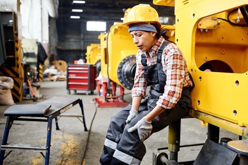 Young female worker of warehouse touching hurting knee while sitting by machine