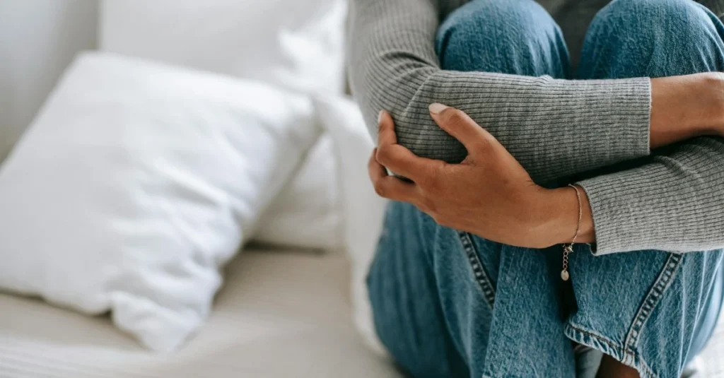 A person in jeans and a grey sweater sits on a bed holding a grey pillow against their stomach, suggesting comfort or relief from abdominal discomfort
