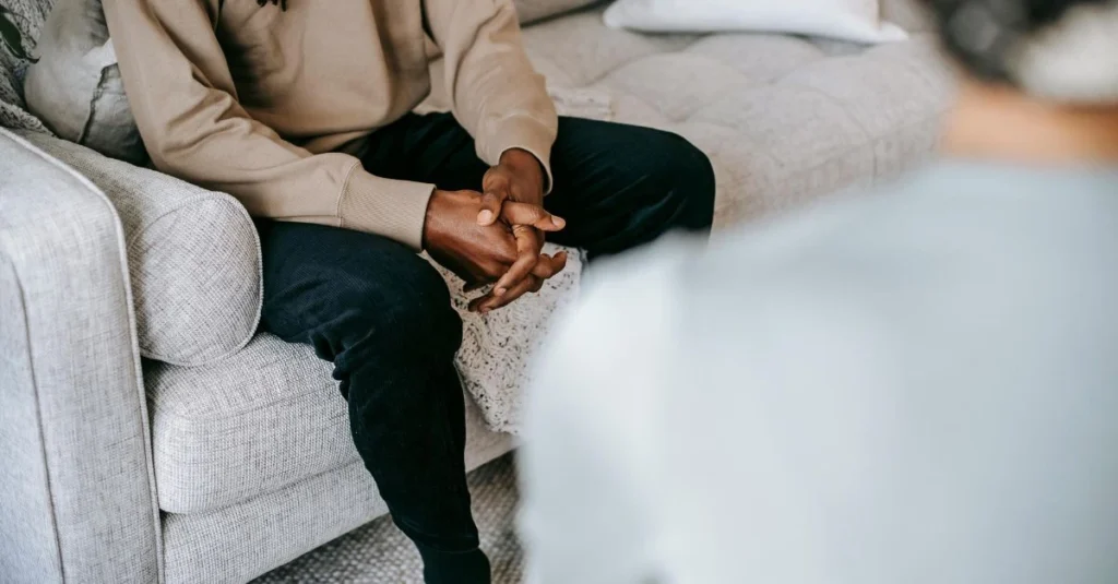 A person in jeans and a grey sweater sits on a bed holding a grey pillow against their stomach, suggesting comfort or relief from abdominal discomfort