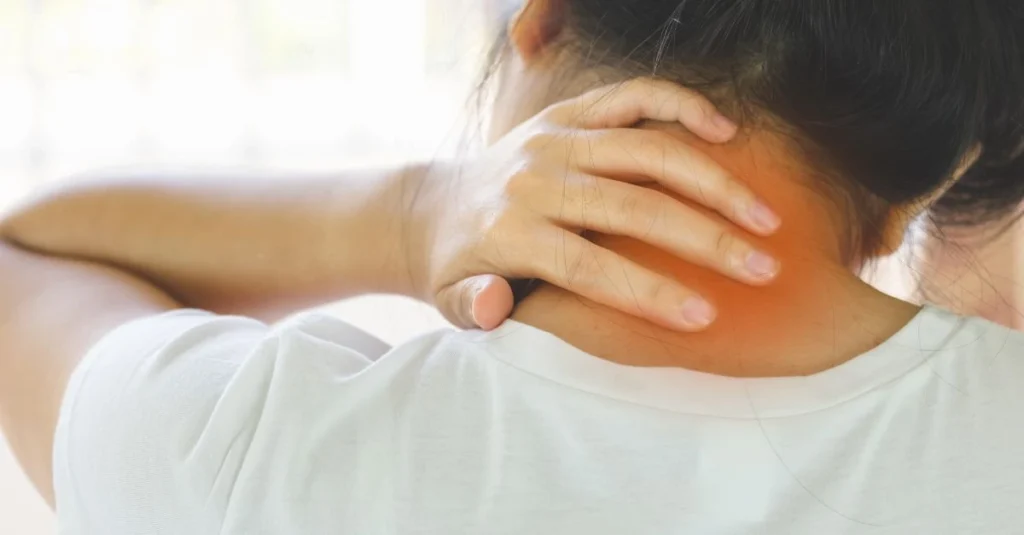 A woman in a white shirt gently holds the back of her neck with her hand, suggesting neck pain or discomfort in soft natural lighting